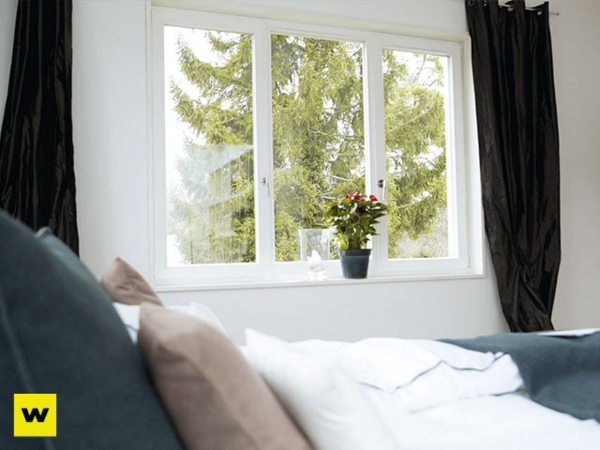Helles Schlafzimmer mit großem Fenster, Blick ins Grüne und Blumentopf auf Fensterbank.
